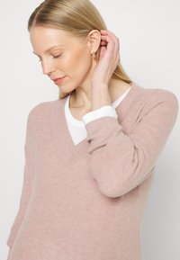 Sweater de malha rosa claro com um decote em V profundo, mangas longas, punhos canelados e uma textura suave. Usado por cima de uma camisa branca.