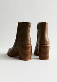Para brązowych skórzanych botków za kostkę z drewnianymi, warstwowymi obcasami, elastycznymi panelami bocznymi i detalem z zamkiem z tyłu, pokazane z tyłu.