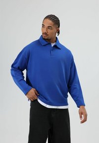 Blauw poloshirt met lange mouwen en een kraag, gemaakt van een zachte, gestructureerde stof. Gedragen over een wit shirt, gecombineerd met donkere broek.