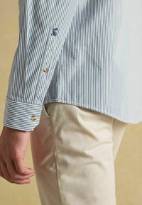 Niebieska i biała w paski koszula z długim rękawem z kontrastowymi mankietami na guziki. Na rękawie znajduje się haftowane logo. Połączona z jasnymi spodniami.