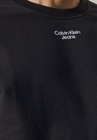 Camiseta negra de cuello redondo con el logo blanco de "Calvin Klein Jeans" impreso en la parte superior izquierda del pecho.