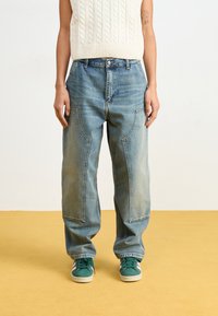 Jeans in denim blu chiaro a gamba larga con tasche oversize, texture sbiadita e cuciture a contrasto; abbinati a scarpe da ginnastica verdi e bianche.