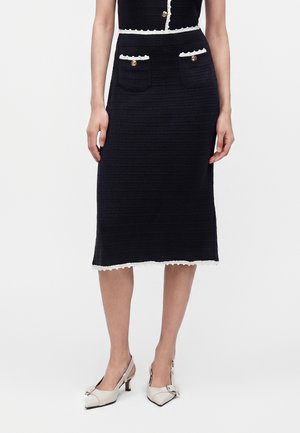 Žena nosi čiernu midi sukňu s bielym lemom a zlatými gombíkmi na vreckách, v kombinácii s bielymi slingback podpätkami so sponami.