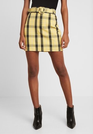 Mini-jupe à carreaux jaunes avec une silhouette ajustée, des accents noirs et une ceinture assortie. Paire de bottines noires avec un bout pointu.