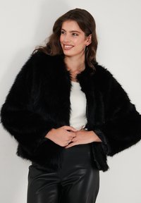 Black faux fur cropped jacket with a soft texture, oversized sleeves, and a rounded collar, paired with a white top and black leather pants.