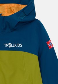 Jachetă de vânt albastră și verde, cu glugă cu fermoar, căptușeală portocalie, logo brodat "TROLLKIDS" și o patch cu steagul norvegian roșu.