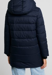 Marinblå uppblåsbar jacka med vadderad design och huva. Har en lång modell och ribbade manschetter. Slät textur med horisontella sömdetaljer.