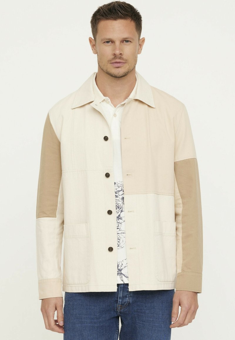 Lichte jas in beige en tan kleuren. Voorzien van een knoopsluiting aan de voorkant, borstzakken en contrasterende mouwpanelen. Textuurstof.