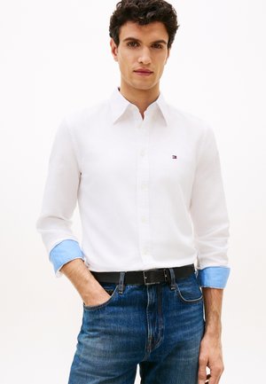 Άνδρας με σγουρά μαλλιά που φοράει λευκό πουκάμισο με μπλε μανσέτες και μικρό λογότυπο, φορεμένο μέσα σε μπλε τζιν παντελόνι με μαύρη ζώνη.