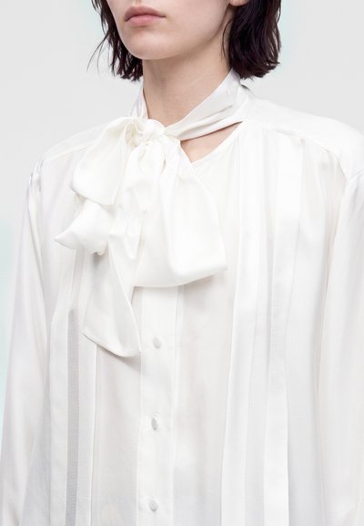 Femme portant un chemisier blanc avec un grand nœud noué au col et des détails plissés verticaux sur le devant.