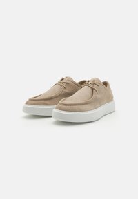 Beige ruskinnsko med en glatt tekstur, som har en hvit gummisåle. Designet inkluderer sydd detaljer og snøring.