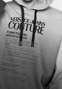 Šedá mikina s kapucí s černým potiskem textu, nastavitelnými šňůrkami, měkkou texturou a pohodlným střihem. Obsahuje specifikace značky a loga.