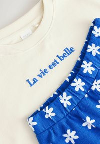 Kremowy sweter z niebieskim haftem "La vie est belle" oraz niebieskie spodenki w kwiaty z białymi wzorami. Miękki materiał, casualowy krój.