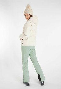 Veste crème rembourrée avec capuche, associée à un pantalon de ski vert clair. Le modèle porte un bonnet en tricot crème avec un détail de pompon.