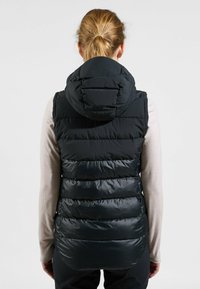 Schwarze wattierte Weste mit Kapuze, ausgestattet mit horizontalen Steppnähten, einer glatten Textur und einem längeren Rückenbereich. Über einem hellen Langarmoberteil getragen.