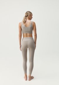 Šviesiai rudos sportinės liemenėlės su plonais dirželiais ir besiūle konstrukcija, derinamos su atitinkančiomis aukštomis tampriomis kelnytėmis, pagamintomis iš lygaus, elastingo medžiagos.