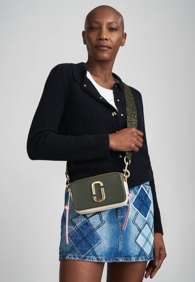 Green leather crossbody bag with gold hardware, pink tassels, and a patterned strap. Paired with a black ribbed cardigan and denim skirt.