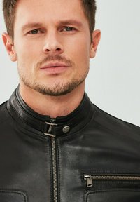 Zwarte leren jas met een opstaande kraag, voorrits en een ritszak op de borst. Glad van textuur met een lichte glans.
