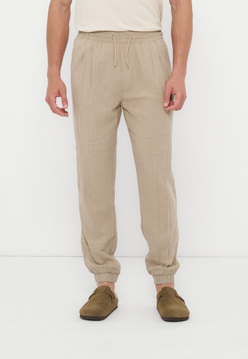 Άνδρας που φοράει μπεζ παντελόνι με κορδόνι και ελαστικές μανσέτες και καφέ παπούτσια χωρίς κορδόνια, στέκεται μπροστά σε λευκό φόντο.