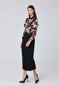 Jupe plissée noire fluide avec une ceinture en cuir ajustée, associée à un chemisier en chiffon floral marron avec des volants. Chaussures à talons noires.