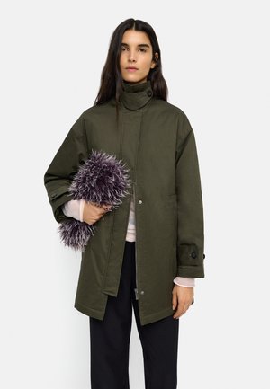 Kobieta ubrana w oliwkowy płaszcz z wysokim kołnierzem i czarne spodnie, trzyma puszystą, fioletowo-białą torebkę kopertówkę o fakturze.