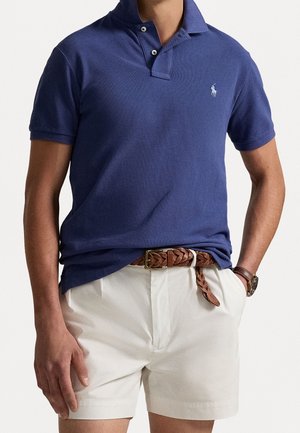 Granatowa koszulka polo z krótkimi rękawami, wykonana z tkaniny o fakturze, z dwoma guzikami przy dekolcie oraz małym haftowanym logo, połączona z białymi szortami i brązowym paskiem.