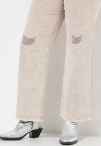 Jeans ampi beige chiaro con toppe strappate alle ginocchia, abbinati a stivaletti argentati lucidi con tacco a blocco.