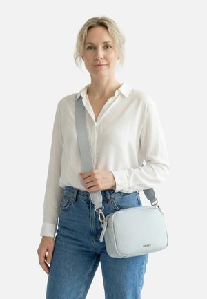 Donna con camicetta bianca e jeans blu che porta una borsa a tracolla grigio chiaro con una tracolla larga sulla spalla.