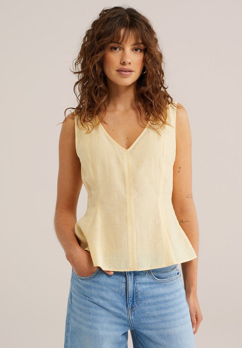 Femme aux cheveux bouclés bruns portant un haut sans manches jaune pâle et un jean bleu clair, debout devant un fond clair uni.