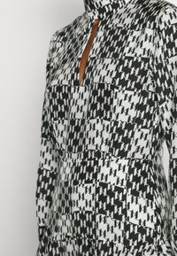 Černobíle vzorované šaty s vysokým límcem, výstřihem ve tvaru klíče a velkými předními kapsami. Tkanina má hladkou texturu.