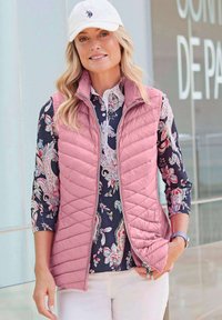 Rosa gesteppte Weste mit hohem Kragen, kombiniert mit einem dunkelblumigen Hemd. Eine weiße Mütze und eine Uhr runden das Outfit ab. Glattes Material, lässiges Design.