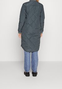 Lång, mörkgrön quiltad kappa med krage, bakre sömmar och långa ärmar, i kombination med ljusblå jeans och svarta plattformsskor.