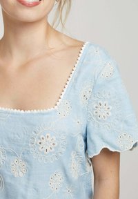 Blouse bleue brodée avec un décolleté carré, des manches à volants et des motifs floraux blancs. Détail délicat le long du décolleté.