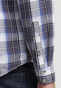 Lyhyet hihansuut, ruudullinen paita sinisessä, harmaassa ja valkoisessa. Puuvillamateriaali, jossa on napit ja ompeluyksityiskohtia hihojen suissa.