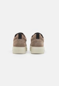 Brązowe zamszowe sneakersy o minimalistycznym designie, z czarną pętelką z tyłu i białą gumową podeszwą. Teksturowana powierzchnia z delikatnym przeszyciem.
