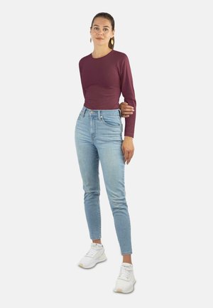 Langärmliges burgunderfarbenes Oberteil kombiniert mit hellblauen hochgeschnittenen Jeans und weißen Sneakers; das Model steht in einer neutralen Haltung.