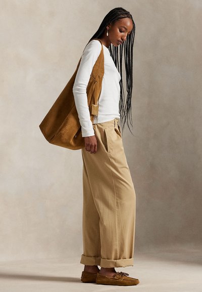 Bruine suède schoudertas, wit shirt met lange mouwen, beige wijde broekspijpen en bruine mocassin-achtige schoenen, tegen een neutrale achtergrond.