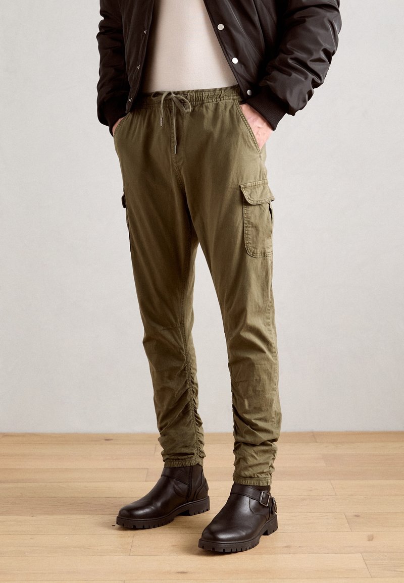 Mannequin portant un pantalon cargo jogger vert olive avec des bords élastiques, associé à des bottines en cuir noir et une veste marron foncé.