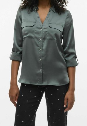 Femme portant une chemise en satin verte boutonnée avec manches retroussées et un pantalon noir à petits pois blancs sur un fond uni.