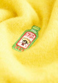 Żółty dzianinowy materiał z haftowaną łatą w kształcie zielonej butelki z napisem "Hot Stuff", na której widnieje osoba w sombrero i czerwonym szaliku.