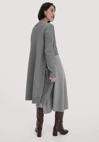 Grauer Wollcardigan mit langen Ärmeln, knielang, mit Seitentaschen und gerippten Akzenten. Getragen über einem passenden grauen Kleid und braunen Absatzstiefeln.