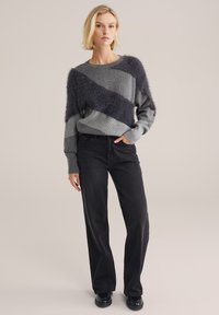 Grauer Pullover mit diagonaler, flauschiger Streifen, gerippten Bündchen, kombiniert mit schwarzen weiten Hosen und schwarzen Schuhen, vor einem neutralen Hintergrund.