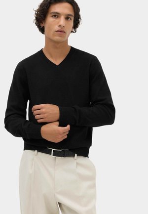 Joven con cabello rizado que lleva un jersey negro de cuello en V y pantalones beige, ajustándose una manga con una expresión facial neutral.