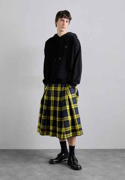 Schwarzer Kapuzenpullover mit Fronttasche, getragen über einem gelben und schwarzen karierten Kilt. Kombiniert mit schwarzen Socken und glänzenden schwarzen Schuhen.
