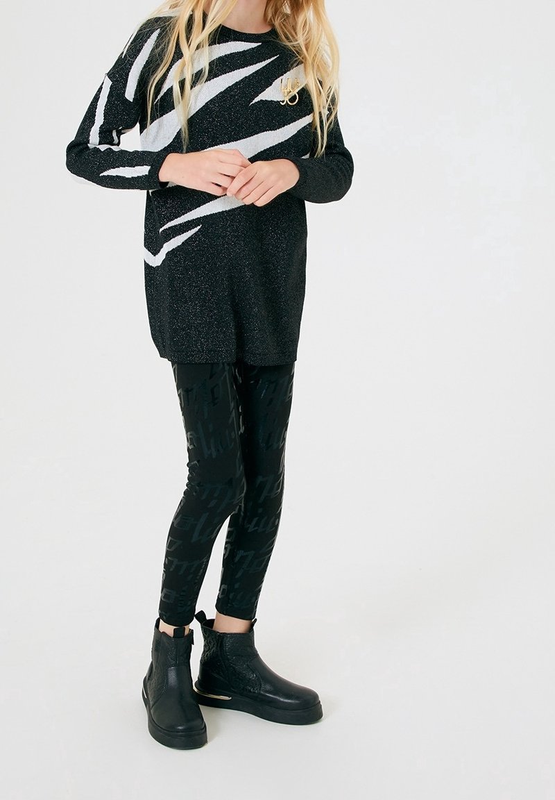 Enfant portant un long pull à motif noir et blanc, des leggings noirs brillants avec impression de texte, et des bottines noires sur fond blanc.