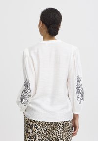Hvid bluse med broderet sort blomsterdesign på ærmerne, løs pasform, tekstureret stof, parret med leopardmønstrede bukser.