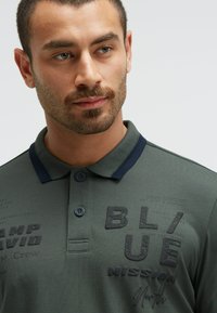 Polo verde con colletto blu navy, lettering testurizzato, chiusura con bottoni e dettagli stampati, realizzato in un materiale liscio e leggero.