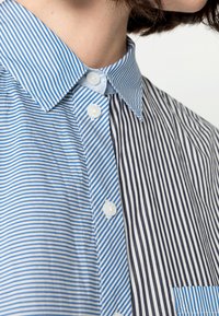 Randig skjorta med ett blått och vitt mönster på ena sidan och mörkblå ränder på den andra. Har en klassisk krage och detaljer med knappar.