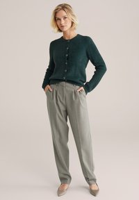 Donkergroen, gebreide cardigan met een ronde hals en zeven gouden knopen. Grijze, hooggewaisted, geplooide broeken en lichtgrijze, spitse flats.