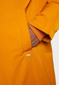 Main d'une personne insérée dans la poche d'une veste orange vif avec une petite étiquette de la marque « Scotch & Soda » sous la poche.
